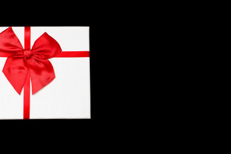 White gift box with a red ribbon and a bow on a black background. Gift for birthday or traditional holiday. Gift box close-up. Free space for text or imageの写真素材