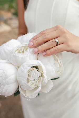 The bride holds a wedding bouquet of white peonies close-up. A beautiful white bridal bouquet of white peonies and the bride's hand with a wedding ring close-up on a vertical photoの写真素材