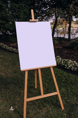 A wooden easel with a white blank sheet is standing outside. An empty easel with a vertical canvas. A clean art easel stands in a garden, trees, flowers and a river in the background. Drawing, artの写真素材