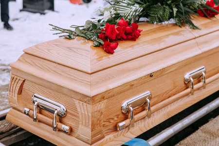 A wooden coffin with flowers on the lid stands above a dug grave in the cemetery. Funeral ceremony at the cemetery in winter. Farewell ceremony and burial.の写真素材