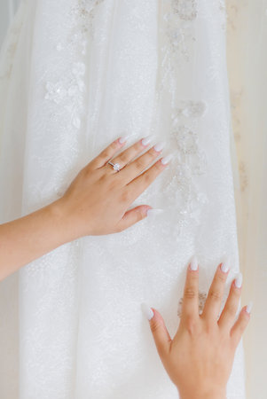 Bride's hands with delicate white manicure on a white wedding dress close-up. Bride touching her dress. Beautiful wedding ring with a precious stone on the bride's fingerの写真素材