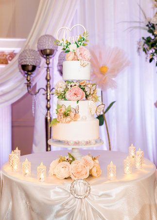 Wedding cake with the love letters on top. Beautiful wedding cake with white cream decorated with flowers and sugar butterflies. Elegant three-tiered wedding cake with candles around it at the banquetの写真素材