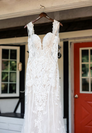 Close-up of a wedding dress hanging on a hanger in front of a house. Lace wedding dress hanging in front of house door outdoors on wedding day for bride, cropped photoの写真素材