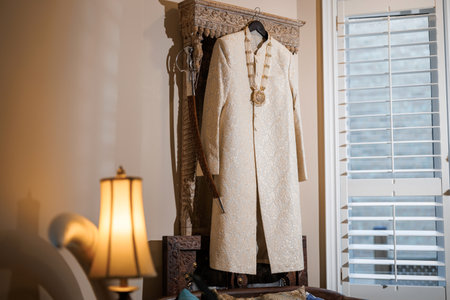 Indian traditional outfit of the groom hangs on a hanger in the room. A festive wedding sherwani with a pattern and a necklace hangs above the bed. Prepared Indian wedding men's wear and Talwar saberの写真素材