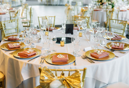Served round tables at a wedding close-up. Table with white tablecloth, gold plates, peach napkins, glasses and silverware for guests. Served table at a celebration without people in a restaurantの写真素材