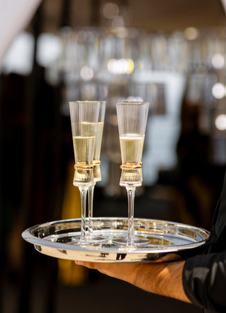 Glasses of champagne on a tray are held by a waiter. Champagne in elegant glasses is served at a banquet. Sparkling wine on a silver tray is served at a wedding. Exquisite alcoholic drink at a partyの写真素材