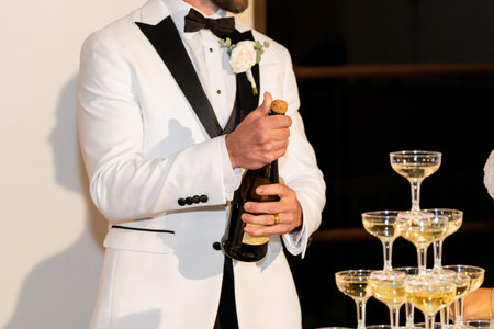 Groom opening champagne on his wedding. A man in a white elegant suit opens a bottle of champagne at a celebration or banquet for a fun and festive mood for the guestsの写真素材