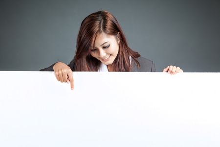 Asian businesswoman stand behind a blank banner , point down and smile on gray backgroundの写真素材