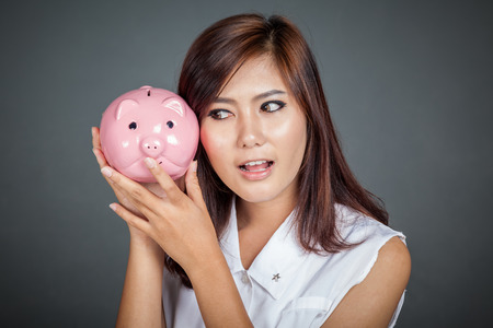 Beautiful Asian girl wonder how much money in pig money box shake it on her ear on gray backgroundの写真素材