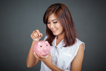 Beautiful Asian girl smile put a coin to pink pig money box on gray backgroundの写真素材