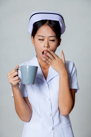 Young Asian nurse yawn with a cup of coffee on gray backgroundの写真素材