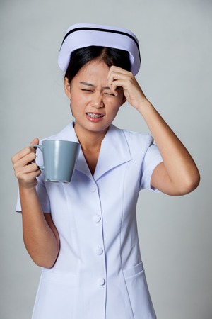 Young Asian nurse got headache with a cup of coffee on gray backgroundの写真素材