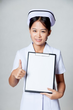 Young Asian nurse thumbs up with blank paper on clipboard on gray backgroundの写真素材
