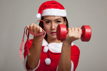 Asian Christmas girl with Santa Claus clothes , measuring tape and dumbbell on gray backgroundの写真素材