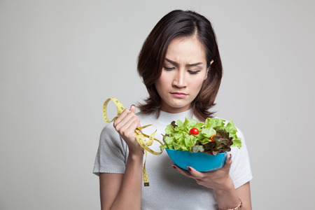 Unhappy Asian woman with measuring tape and salad on gray backgroundの写真素材
