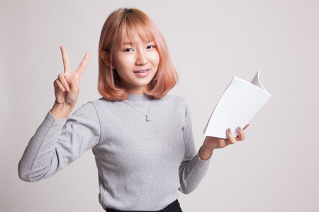Young Asian woman show victor sign with a book on gray backgroundの写真素材