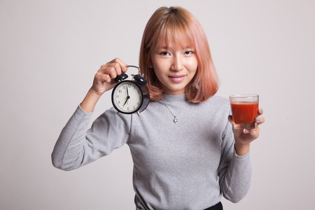 Young Asian woman with tomato juice and clock on gray background