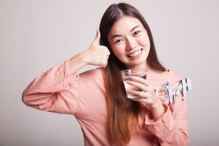 Young Asian woman thumbs up with a glass of drinking water on gray backgroundの写真素材