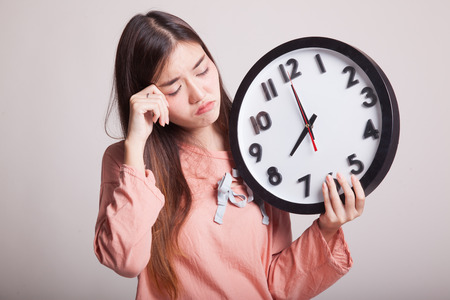 Sleepy young Asian woman with a clock in the morning on gray background