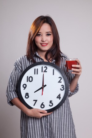 Young Asian woman with tomato juice and clock on gray background