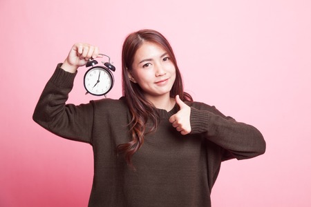 Young Asian woman show thumbs up with a clock on pink backgroundの写真素材