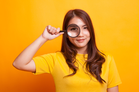 Young Asian woman with a magnifying glass in yellow dress on yellow backgroundの写真素材