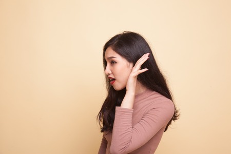 Beautiful young Asian woman listen to something on beige background