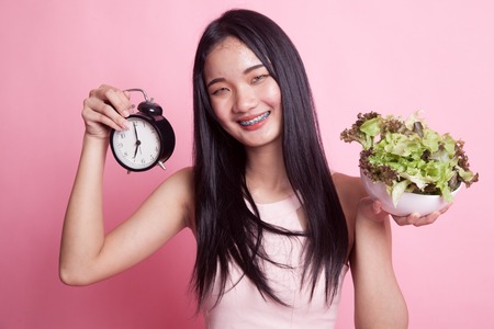 Young Asian woman with clock and salad on pink backgroundの写真素材