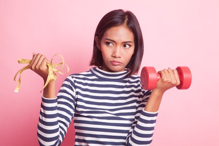 Exhausted  Asian woman with dumbbells and measuring tape on pink backgroundの写真素材