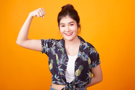 Beautiful young Asian woman flex bicep in  tropical shirt on yellow backgroundの写真素材