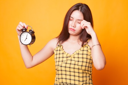 Sleepy young Asian woman with a clock in the morning on yellow background