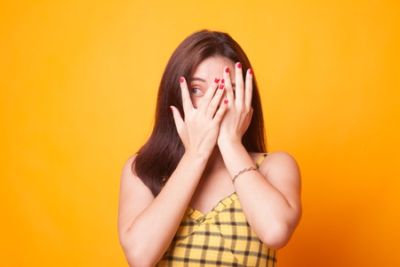 Beautiful young Asian woman peeking through fingers on yellow backgroundの写真素材