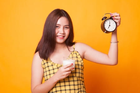 Healthy Asian woman drinking  glass of milk hold clock on yellow backgroundの写真素材