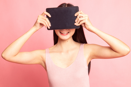 Young Asian woman with a computer tablet over her face on pink backgroundの写真素材