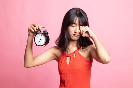 Sleepy young Asian woman with a clock in the morning on pink background