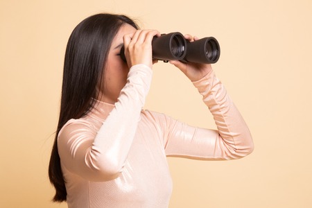 Young Asian woman with binoculars on  beige background