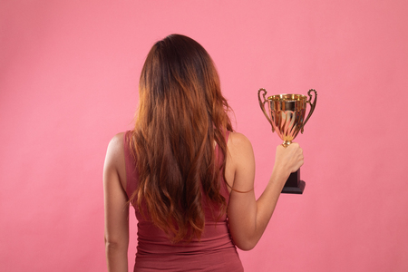 Back view of successful young asian woman holding a trophy on pink backgroundの写真素材
