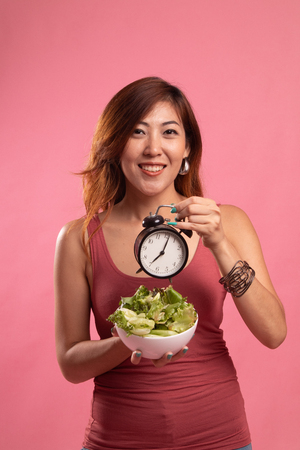 Young Asian woman with clock and salad on pink backgroundの写真素材