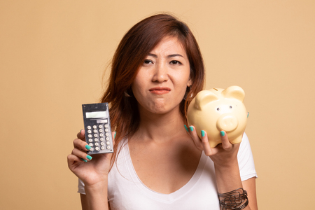 Unhappy Asian woman with calculator and piggy bank on beige backgroundの写真素材