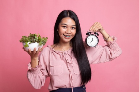 Young Asian woman with clock and salad on pink background