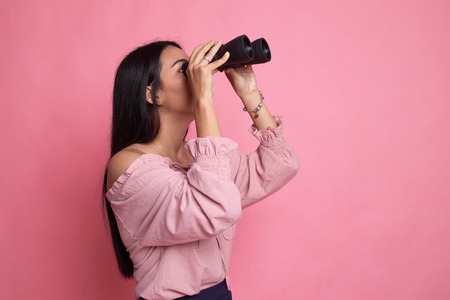 Young Asian woman with binoculars on pink background