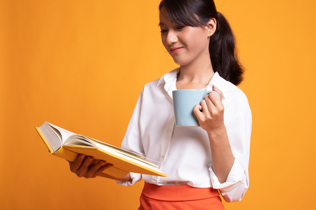Young Asian woman with a book and cup of coffee on yellow backgroundの写真素材