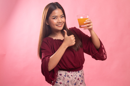 Young Asian woman thumbs up drink orange juice on pink backgroundの写真素材