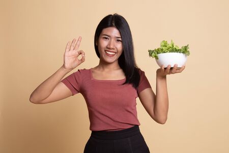 Healthy Asian woman show OK with salad on beige background
