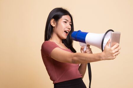 Angry  young Asian woman shout with megaphone to mobile phone on beige backgroundの写真素材