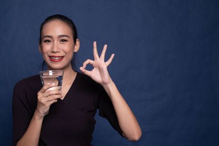 Young Asian woman show OK with a glass of drinking water on blue backgroundの写真素材
