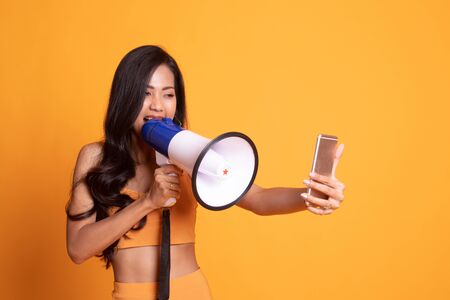 Angry  young Asian woman shout with megaphone to mobile phone on yellow backgroundの写真素材