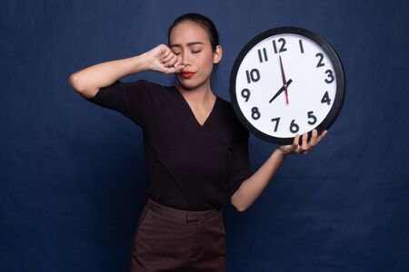Sleepy young Asian woman with a clock in the morning on blue background