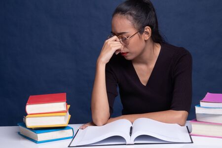 Exhausted Asian woman got headache read a book with books on table on blue background