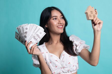 Happy young asian woman with house model and bunch of money banknotes on cyan color backgroundの写真素材